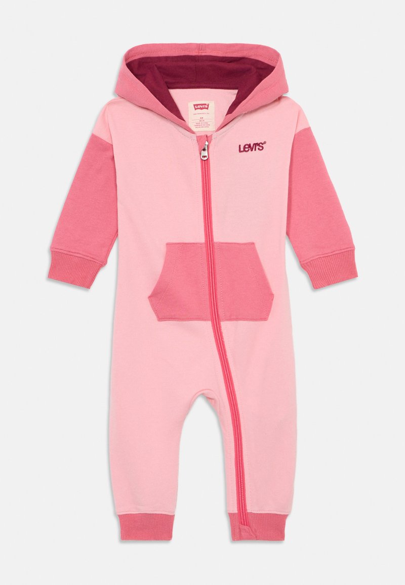 Combinaison zippée rose bébé avec capuche, présentant des manches d'un rose plus foncé en contraste, une poche avant et des poignets côtelés. Fabriquée en tissu doux.