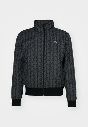 Schwarze Reißverschlussjacke mit geometrischem Muster, hohem Kragen, gerippten Bündchen und Saum sowie kleinem grünen Lacoste-Logo auf der linken Brust.