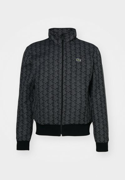 Veste zippée noire présentant un motif géométrique, un col montant, des poignets et un ourlet côtelés, avec un petit logo vert Lacoste sur la poitrine gauche.