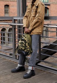 Kurtka użytkowa koloru khaki z licznymi kieszeniami, plecak w kamuflażowym wzorze z zielonymi i czarnymi akcentami, szare jeansy i czarne sznurowane buty.