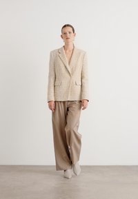 Beige tweed-blazer med to frontlommer, paret med løse tan bukser og lyse ankelstøvler, satt mot en enkel bakgrunn.