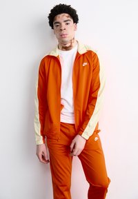 Nike αθλητική φόρμα σε πορτοκαλί χρώμα με κίτρινες λεπτομέρειες, περιλαμβάνοντας μπουφάν με φερμουάρ και αντίστοιχες παντελόνες. Λείος υφασμάτινος τύπος, με μινιμαλιστική λογότυπο στο στήθος.
