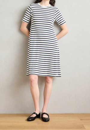 Femme portant une robe blanche avec des rayures horizontales noires, manches courtes, ballerines noires à bride à boucle, debout contre un mur beige et un sol en bois.