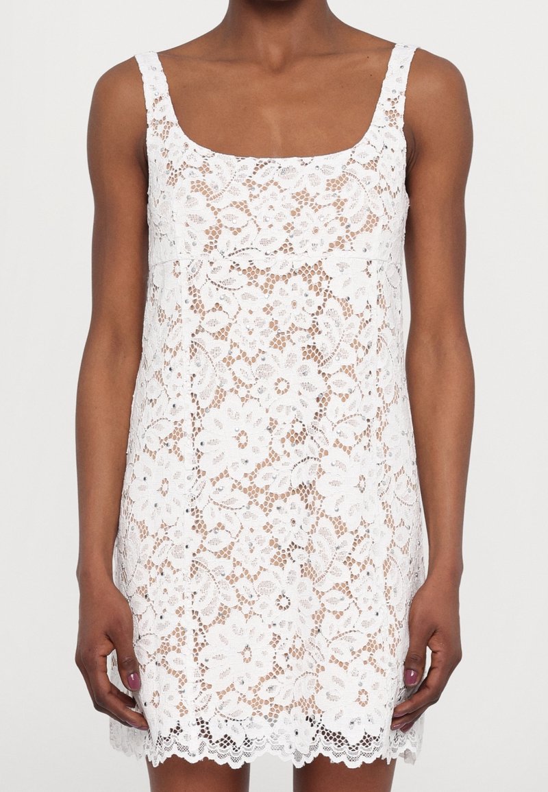 Femme portant une mini-robe blanche sans manches avec une superposition en dentelle florale et un ourlet festonné, montrant la peau bronzée à travers le motif de dentelle.