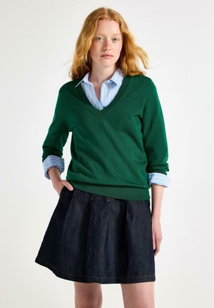 Ung kvinde med rødt hår, iført en grøn V-hals sweater over en blå stribet skjorte og en mørk denimnederdel, stående med den ene hånd i lommen.