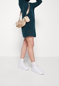 Béžová kabelka přes rameno s řetízkovými detaily a logo popruhem, nošena přes tyrkysové pletené šaty. Model má na sobě bílé tenisky s kotníkovými ponožkami.