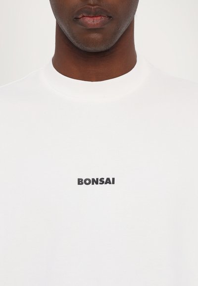 T-shirt en coton blanc avec un col rond et le texte noir "BONSAI" imprimé sur la poitrine. Texture lisse, coupe classique.