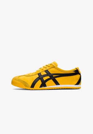 Gele sportieve schoen met zwarte accenten, gladde leren textuur, gebogen ontwerp, ronde teen, platte witte zool en opvallende branding aan de zijkant.