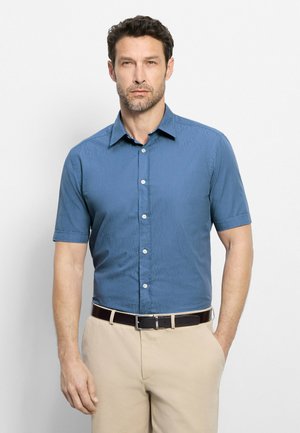 Uomo con capelli corti e scuri e barba, che indossa una camicia blu a maniche corte e pantaloni beige con cintura nera, in piedi davanti a uno sfondo bianco.