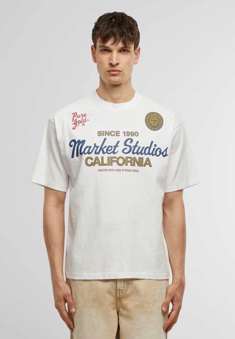 Balts kokvilnas T-krekls ar zilo un zeltaino tekstu: "Market Studios CALIFORNIA", ar sarkaniem akcents un logotipa detaļām uz krūtīm.