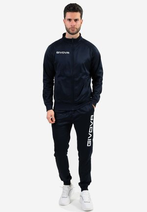 Tuta da ginnastica blu navy realizzata in tessuto liscio, con colletto alto e zip frontale. Logo "GIVOVA" in bianco su maglia e pantaloni. Indossata con sneakers bianche.