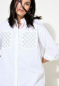 Camicia bianca con bottoni e due tasche decorative adornate da borchie argentate disposte a motivo a diamante. Vestibilità ampia con maniche arrotolate.