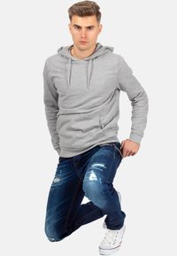 Felpa grigia con coulisse, tasca frontale; jeans blu strappati. Modello in ginocchio, indossa sneakers bianche, sfondo neutro.