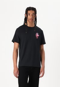 Sort bomulds T-shirt med et pink grafik på venstre bryst. Designet inkluderer stjerner og palmetræer. Korte ærmer, rund halsudskæring.