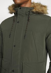 Parka di colore verde oliva realizzata in tessuto liscio, con cappuccio foderato in pelliccia, lacci a contrasto, bottoni a pressione e grandi tasche anteriori.