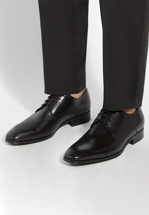 Chaussures formelles en cuir noir poli à lacets, portées avec un pantalon foncé sur un fond clair.