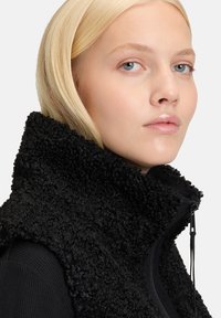 Czarna polarowa kamizelka z wysokim kołnierzem, teksturowany materiał, z zapięciem na zamek z przodu. Gładka podszewka widoczna na krawędzi kołnierza, minimalne okucia.