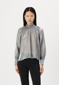 Blusa argento trasparente con collo alto, design plissettato, maniche lunghe e polsini elasticizzati. Abbinata a pantaloni neri aderenti e scintillanti.