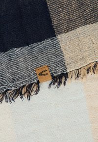 Vevet stof med marineblå, beige og cremefarvet ternet mønster og frynser på kanterne, der har et lille brunt mærke med et sort logo.