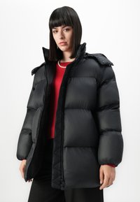Vrouw met steil zwart haar, gekleed in een zwarte pufferjack, rode top, parelketting en zwarte broek tegen een witte achtergrond.