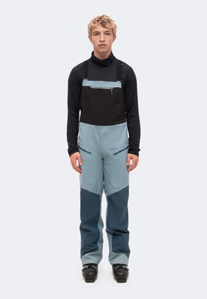 Un homme présente un pantalon de ski à bretelles isolant, bicolore bleu et noir. Il porte un haut noir et des chaussures de ski, sur un fond uni.