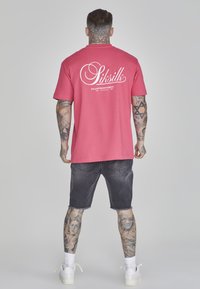 Camiseta de algodón rosa con el logo blanco de 'Siksilk' en la parte de atrás, combinada con pantalones cortos de mezclilla oscura y zapatillas blancas. Figura masculina tatuada de espaldas.