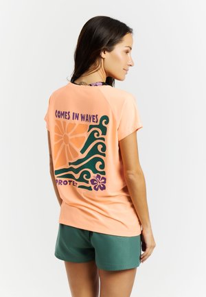 Vrouw die naar rechts kijkt, draagt een perzikkleurig T-shirt met golf- en bloemontwerp en de tekst "Comes in waves", gecombineerd met groene shorts tegen een witte achtergrond.