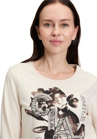Beige Langarmshirt mit floralem und textuellem Druck in Braun und Kristallakzenten, Rundhalsausschnitt und weicher Stoffstruktur.