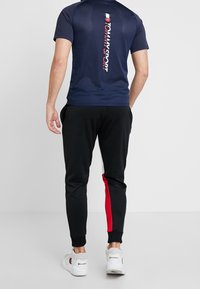 Homme portant une chemise à manches courtes bleu marine avec le logo vertical "Tommy Sport", un pantalon de jogging noir avec une bande rouge, et des baskets blanches Champion, de dos.