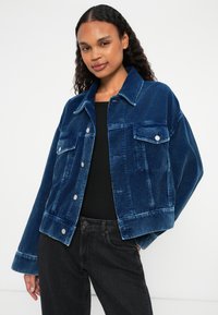 Giacca in corduroy blu con colletto, tasche frontali, chiusura con bottoni e maniche ampie, abbinata a una maglia nera e jeans scuri.