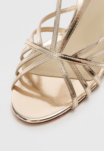 Strappy high-heeled sandal in metallic gold leather; features crisscross straps, a rounded toe, and a glossy finish. Smooth inner lining.