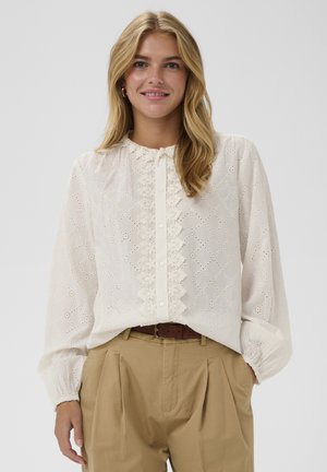 Femme blonde souriante portant un chemisier blanc en dentelle à manches longues avec des boutons à l'avant et un pantalon plissé beige taille haute avec une ceinture marron.