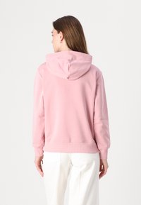 Frau mit langen braunen Haaren, die einen hellrosa Hoodie und weiße Hose trägt, von hinten vor einem einfarbigen weißen Hintergrund.
