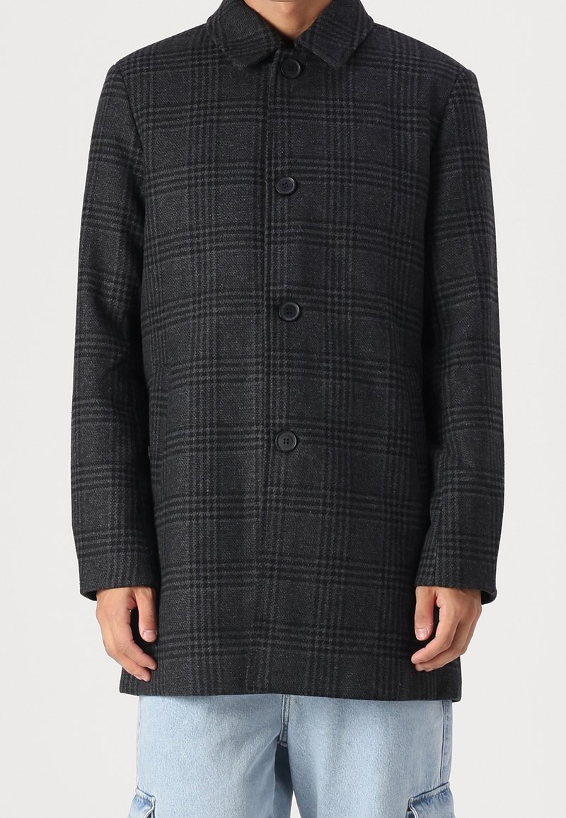 Hombre vistiendo un abrigo de cuadros gris oscuro abotonado con cuello y pantalones de mezclilla azul claro frente a un fondo simple.