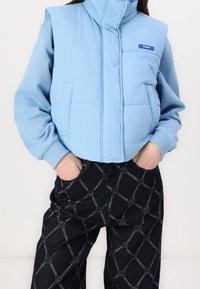 Helles blaues, plüschiges Steppweste mit hohem Kragen und Fronttaschen, kombiniert mit dunkel gemusterten Jeans, die ein kettendesign in Grau aufweisen.