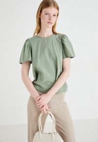 Svetlo zelena kratkodlaka majica s okruglim izrezom i napuhanim rukavima, uparena s bež hlačama. Majica ima suptilno teksturiranu tkaninu.