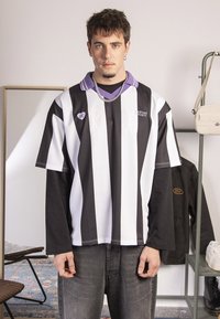 Cămașă supradimensionată cu dungi orizontale alb-negru, cu guler mov, detalii cu logo și o croială lejeră, purtată cu blugi gri.