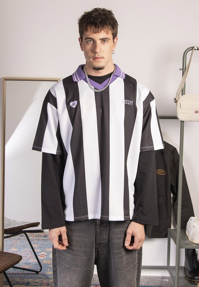 Cămașă supradimensionată cu dungi orizontale alb-negru, cu guler mov, detalii cu logo și o croială lejeră, purtată cu blugi gri.