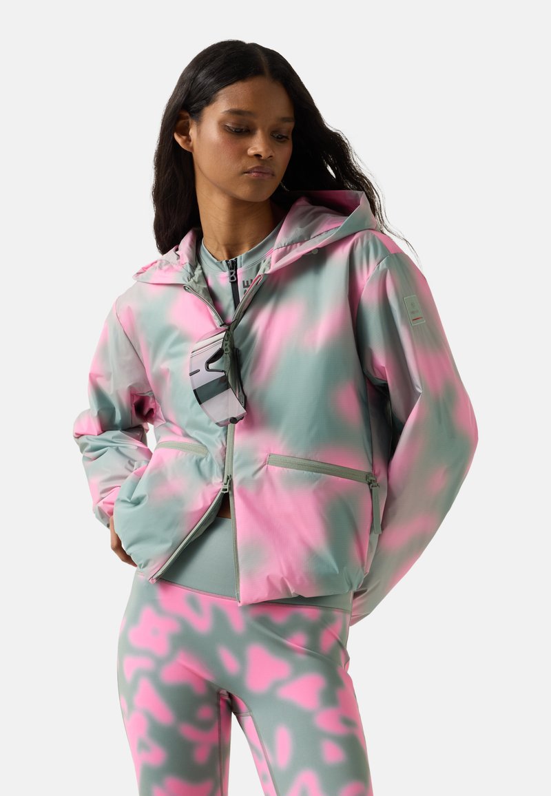 Kvinde iført pink og grå camouflage-mønstret jakke og leggings med hænderne i jakkelommerne, kigger til siden.