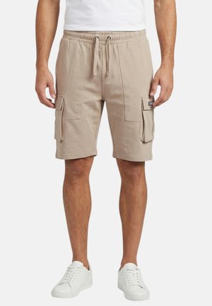 Homme portant un short cargo beige à taille coulissante avec poches latérales, des baskets blanches et un t-shirt blanc, debout sur un fond uni.