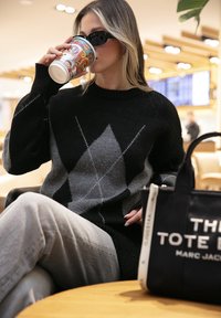 Vrouw in een zwart-grijze ruittrui en zonnebril drinkt uit een kleurrijke mok terwijl ze met gekruiste benen in een café zit.