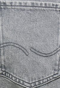 Il tessuto di denim grigio presenta una consistenza liscia con una leggera sbiaditura. Cuciture decorative creano due linee curve sopra una tasca.