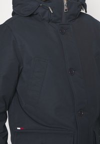 Laivastonsininen eristetty takki hupulla, jossa on nappikiinnitys, kaksi etutaskua ja hillitty punavalkoraidallinen koriste.