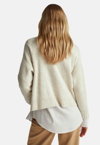 Béžový pletený svetr s kleslými rameny a žebrovaným lemem, přesažený přes bílou pruhovanou košili s límečkem, nošený s hnědými kalhotami.