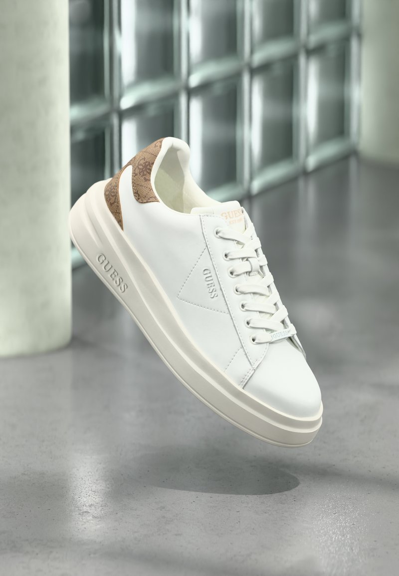 Weiße Low-Top Guess Sneaker mit tanfarbenem gemustertem Fersenbereich, geprägtem Markennamen an der Seite und Sohle, schwebend über einem glänzenden Betonboden.
