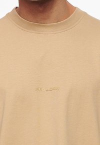 Beige bomulds t-shirt med rund hals og korte ærmer, med prægede "PEGADOR" logo foran. Glat tekstur og afslappet pasform.