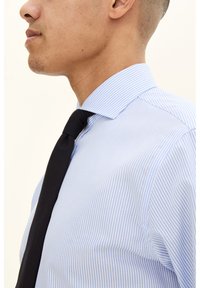 Vaaleansininen, raidallinen paitapusero, jossa on tavallinen kaulus, yhdistettynä mustaan solmioon. Kangas näyttää sileältä ja siinä on hienovarainen kiilto.