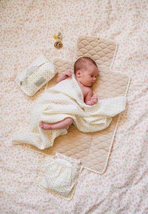 Bebé recién nacido envuelto en una manta blanca, acostado sobre un colchón cambiador acolchado beige sobre una tela con estampado floral, con toallitas para bebés y chupetes cerca.