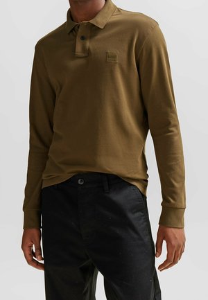 Olijfgroene polo met lange mouwen en een kraag, voorzien van een knoopsluiting en een vierkante logo-patch op de linkerkant van de borst. Gecombineerd met donkere broeken.