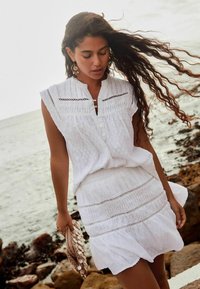 Robe blanche texturée avec une taille cintrée, un ourlet à volants et des manches courtes. Elle présente des détails en boutons et un bord en dentelle. Pochette tissée avec des coquillages.